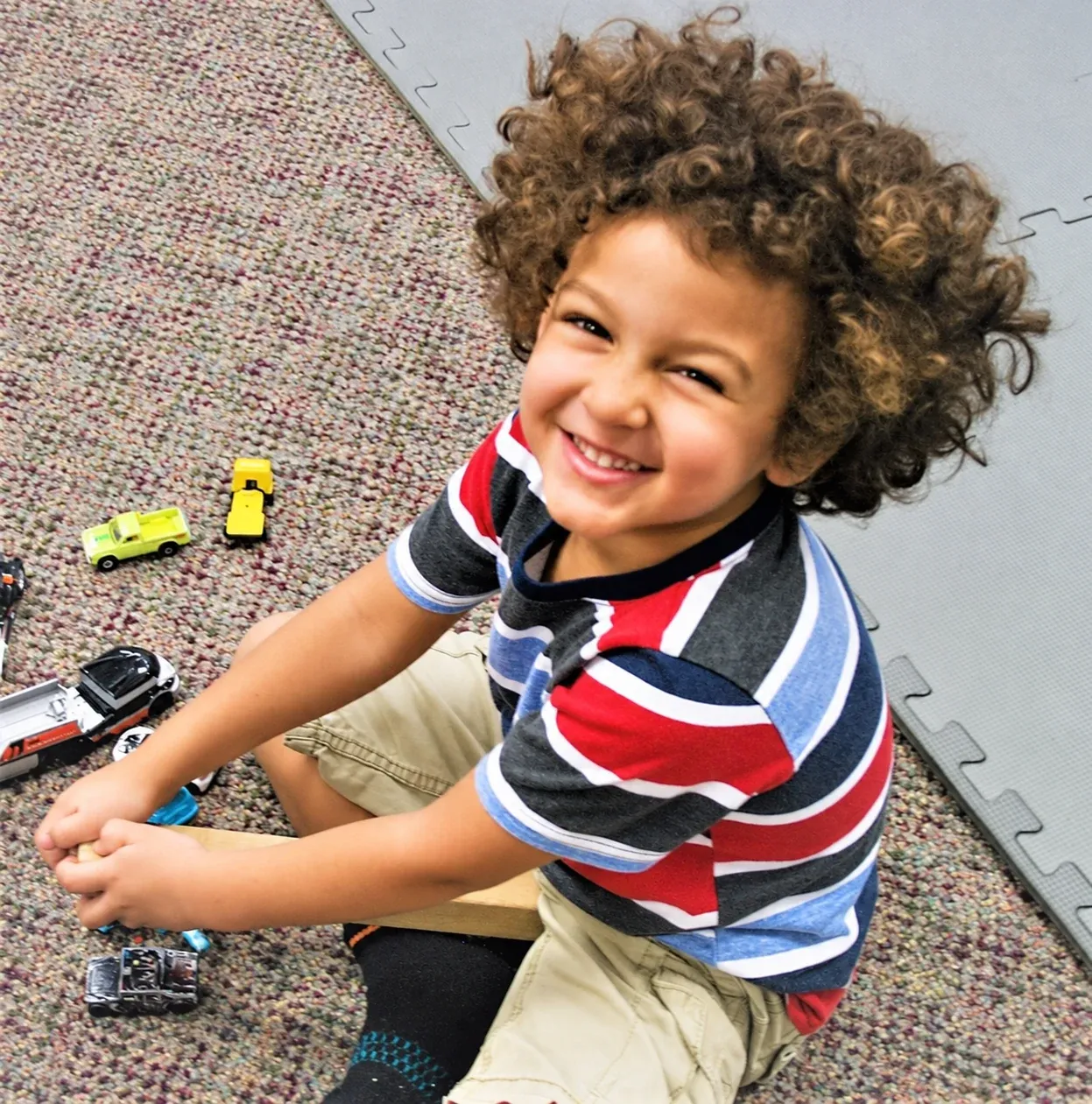 A curly-haired boy grins while playing with toy cars on the classroom floor