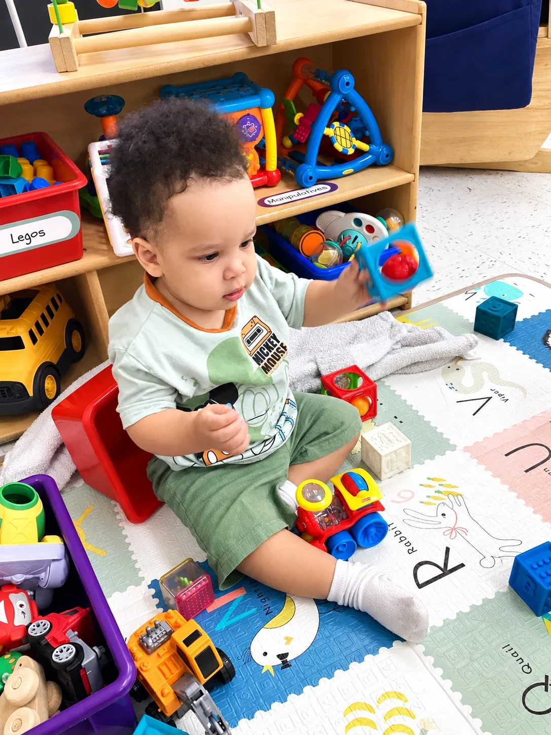 A toddler sits on the alphabet mat surrounded by blocks and toy trucks