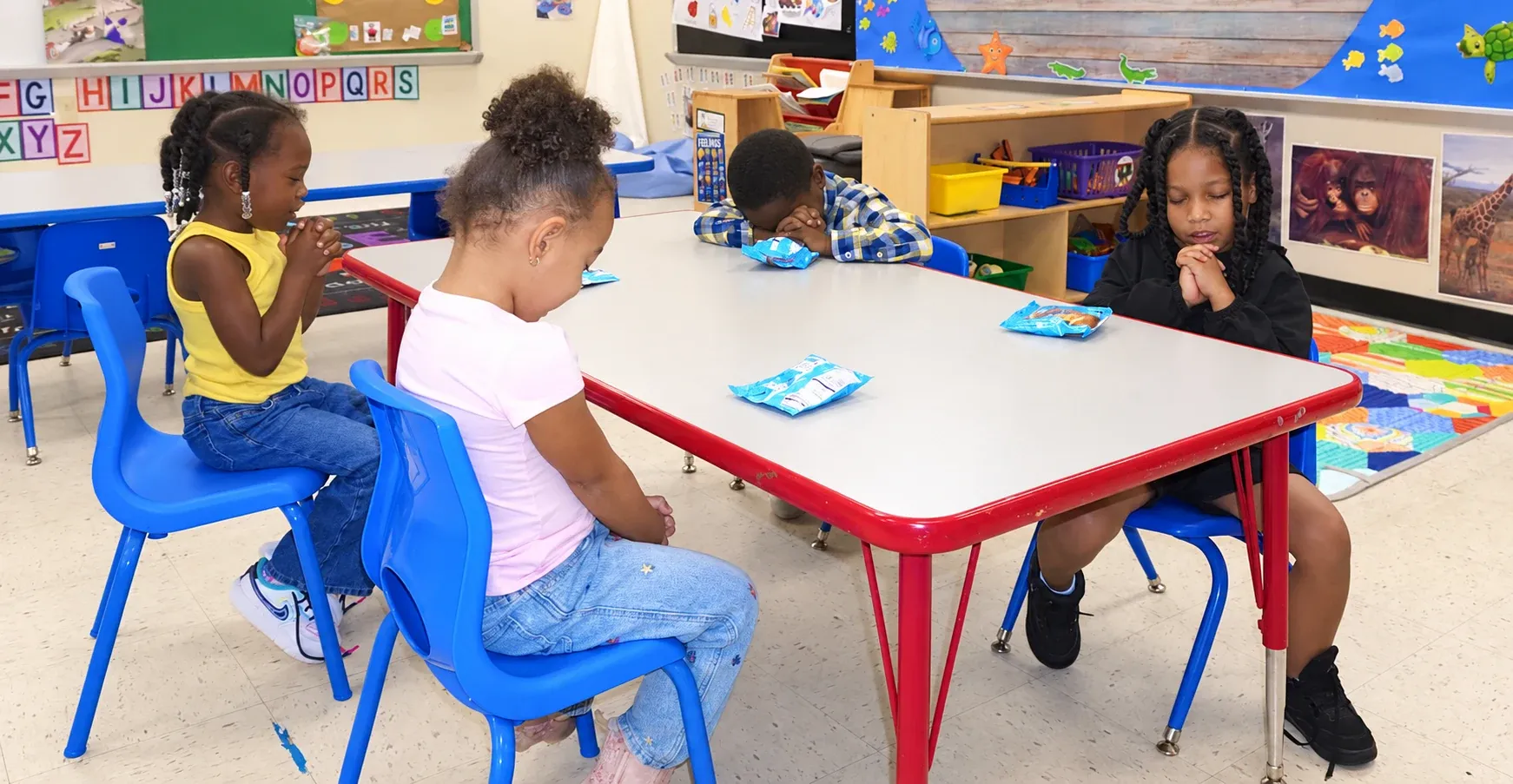 Four preschoolers bow their heads to pray before snack time at their classroom table
