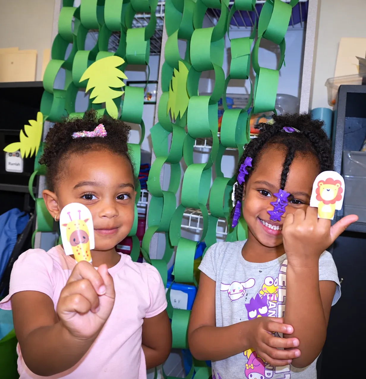 Two girls show off their finger puppets in front of a green paper-chain wall